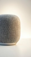 Cylindrical smart speaker with fabric mesh covering in neutral beige tone and white base, positioned on white surface against soft gradient background for tech product display.