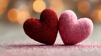 Two textured hearts in burgundy and pink tones touching together on glittery surface with warm bokeh lights creating romantic Valentine's Day atmosphere.