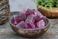 Raw purple amethyst crystals in rustic wooden bowl on weathered table surface, with natural decorative elements in soft background lighting.