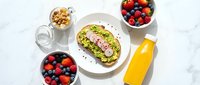 Healthy breakfast spread with avocado toast topped with seeds, fresh mixed berries in bowls, granola, milk and orange juice on white marble surface.