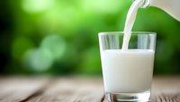 Fresh milk pouring from bottle into clear glass on wooden surface against blurred green natural background, capturing dynamic flow and freshness.