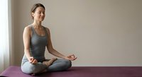 Young Caucasian woman practicing yoga meditation in lotus pose on purple mat at home wearing gray athletic wear in peaceful indoor setting.