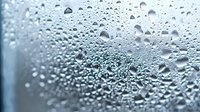 Water droplets and condensation on glass surface create natural abstract pattern with soft blue tint background, macro photography showing crystal clear drops.