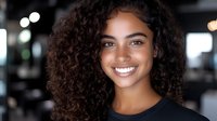 Young mixed-race woman with curly hair smiling confidently in modern office environment. Professional headshot for business and career content.
