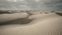 Vast desert landscape with rolling sand dunes under dramatic cloudy sky. Lone figure walking across endless sandy terrain creating sense of scale and solitude.
