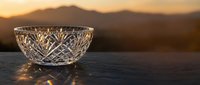 Crystal cut glass bowl on dark surface with golden sunset mountain backdrop, light refracting through intricate facets creating warm glowing reflections.