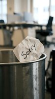 Crumpled paper note with word Sorry tossed in metal trash can in blurred office, symbolizing rejected apology, regret, or failed communication.