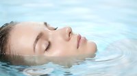 Young woman with closed eyes floating peacefully in clear blue water, face partially submerged showing serene expression for spa and wellness concepts.
