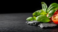 Fresh basil leaves and sliced cherry tomatoes arranged with dark stones on black slate background, dramatic lighting creates contrast for food photography.