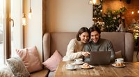 Happy young Caucasian couple sitting in cozy cafe, smiling and looking at laptop screen together, coffee cups on table.
