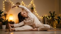 Young woman practicing yoga side stretch pose in cozy bedroom with warm fairy lights creating peaceful evening atmosphere for wellness and relaxation routine.