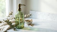 Green glass soap dispenser with eucalyptus branches on marble bathroom counter near window creating spa-like atmosphere for wellness and self-care routines.