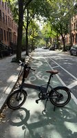 Folding bicycle parked on tree-lined urban street with bike lane, surrounded by brownstone buildings and dappled sunlight, ideal for transportation and city lifestyle content.
