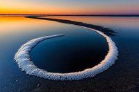 Circular salt formation on dark beach at golden hour sunset with calm water reflections and dramatic sky colors creating natural mineral ring pattern.