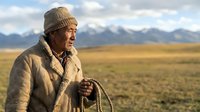 Elderly Asian man in traditional clothing holding rope in vast mountain steppe landscape. Nomadic herder portrait with dramatic sky and golden grassland backdrop.