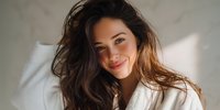 Young brunette woman with long wavy hair smiling warmly while lying in white bedding, natural morning light creating soft shadows for lifestyle content.