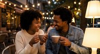 Happy African American couple enjoying coffee together on romantic date night with warm string lights creating cozy intimate atmosphere at outdoor cafe.