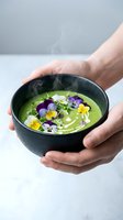 Hands holding black bowl with steaming green soup garnished with edible flowers and microgreens, perfect for fine dining and culinary content.