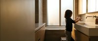 Young child reaching for bathroom sink faucet in modern home interior with natural window light creating warm atmosphere for daily hygiene routine.