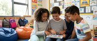 Three diverse elementary school children collaborating on tablet computer in colorful classroom with educational posters and bean bag seating area.