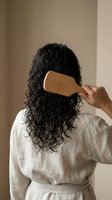 Woman with curly dark hair brushing with wooden comb in bathroom wearing white robe for natural hair care routine and wellness lifestyle.