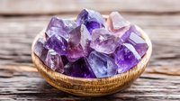 Raw amethyst crystal chunks in wooden bowl on rustic table, purple quartz mineral specimens displaying natural geometric formations and color variations.