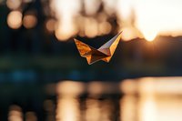 Orange paper butterfly floating against blurred sunset background with bokeh lights and water reflections, creating dreamy atmospheric scene at golden hour.