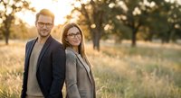Professional business couple in suits standing back to back outdoors during golden hour sunset, representing teamwork and partnership in corporate environment.