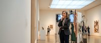 Woman in professional black blazer standing in modern art gallery space with contemporary artwork and sculptures displayed on walls and pedestals, suitable for business, culture, and creative industry concepts.
