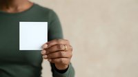 Woman with dark skin tone wearing a green long-sleeve shirt holding a blank white card or paper in front of her chest against a neutral beige background for mockup and branding purposes.