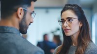 Young woman with glasses in serious conversation with male colleague in office environment. Professional workplace interaction showing attentive listening and engagement.