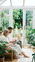 Happy couple working on laptops in bright sunroom with plants while children play in garden outside through glass doors for remote work lifestyle.