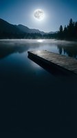 Wooden dock extending into calm lake under full moon with misty mountains and evergreen forest at night, suitable for nature and landscape backgrounds.