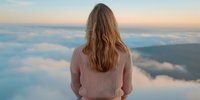 Young woman in pink sweater standing on mountain peak above clouds during sunset, view from behind, contemplating scenic panoramic vista of misty horizon.