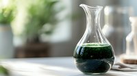 Clear glass carafe with emerald green liquid against blurred background, dramatic lighting creates elegant scientific or mixology atmosphere.