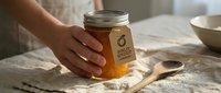 Unrecognizable person holding jar of golden honey on rustic linen surface with wooden spoon, natural lighting for food and culinary content.
