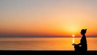 Woman practicing yoga meditation at sunset by calm ocean water with golden sky reflection creating peaceful mindfulness moment for wellness and relaxation.