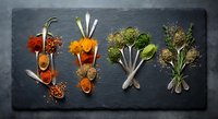 Colorful spices and fresh herbs arranged on dark slate background with wooden spoons creating artistic culinary composition for cooking and seasoning.