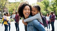 Happy African American mother and daughter embracing outdoors in park setting with diverse families in background during community gathering or school event.