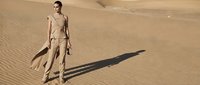 Young adult male, Middle Eastern or North African ethnicity, wearing tan/beige tactical or military-style clothing, holding a surfboard in vast desert landscape with rippled sand patterns and dramatic shadow.
