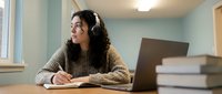 Young woman with long dark curly hair and headphones studying at desk with laptop, notebook and books in bright room for online learning and education.