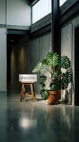Modern industrial office lobby with a welcome sign on a wooden stool beside a lush monstera plant, concrete walls and polished floor.