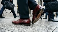 Business professional walking in brown leather dress shoes on city street during rush hour commute with blurred pedestrians in background.