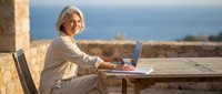 Senior woman with gray hair working on laptop computer outdoors at wooden table during golden hour sunset, remote work lifestyle concept.