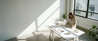 Woman with medium skin tone, wearing beige cardigan, sitting at white desk in bright minimalist office space with natural sunlight, working on laptop surrounded by plants and papers.