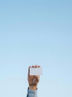 Arm of male person with medium skin tone holding blank white card against clear blue sky, suitable for business messaging, promotional campaigns, and advertising mockups.