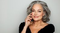 Confident mature woman with silver gray hair smiling warmly while touching face, representing aging gracefully and self-care for skincare and beauty campaigns.
