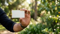 Close-up of male hand holding blank white card outdoors in natural garden setting with blurred green foliage background for business card mockup.