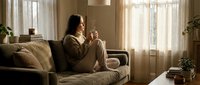 Young woman with dark curly hair relaxing on beige sofa in cozy living room, holding warm beverage cup. Soft natural lighting creates peaceful morning atmosphere.