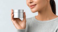 Young Asian woman holding silver cosmetic cream jar against light background, showing natural smile and wearing casual gray sweater. Perfect for beauty and skincare advertising.